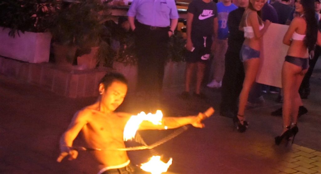 A street performer in Bangkok red light district juggles fire sticks while two sex workers turn and smile Photo Credit: Borderless News Online